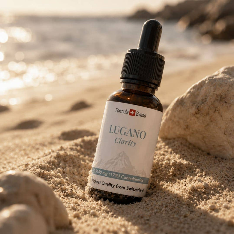 Formula Swiss Lugano Clarity CBD olje i en flaske, presentert i strandsand ved siden av steiner og solbelyst vann.